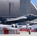 An F-35A Lightning II fighter jet bearing 'AK' tail code and serial number 15534 is captured during takeoff or low-altitude flight at what appears to be an airshow. The aircraft is in a dark gray stealth finish with landing gear still deployed. A crowd of spectators and military hangars are visible in the background.