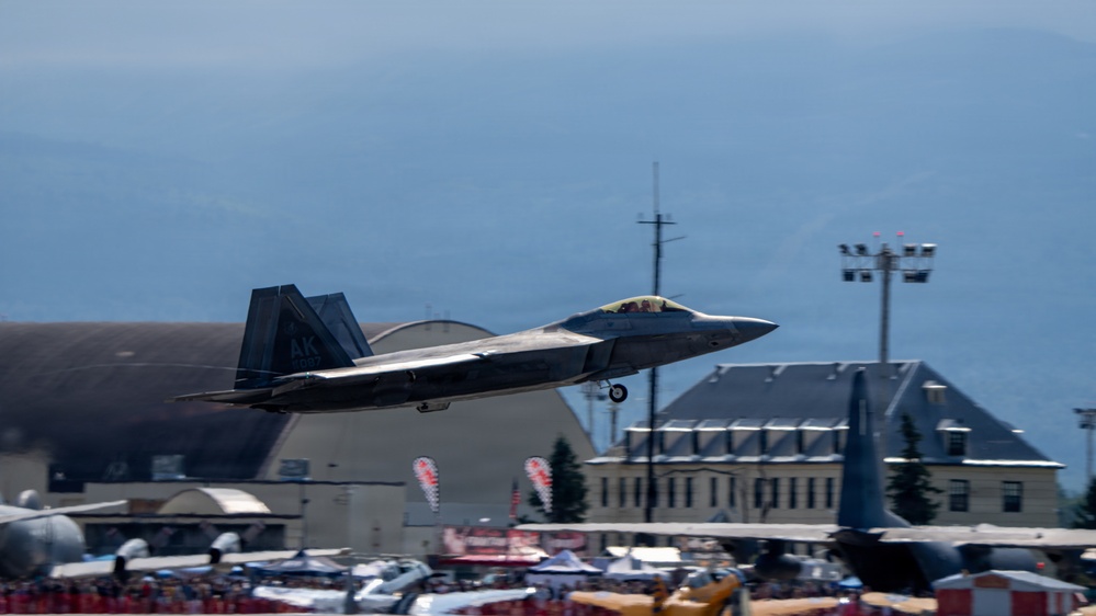 An F-22 Raptor stealth fighter jet marked with 'AK' tail code is captured in low-altitude flight during what appears to be an airshow. The aircraft is flying over a crowded airfield with spectators, hangars, and military base buildings visible in the background. Mountains or hills are visible in the distant background under a hazy sky.