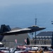 An F-22 Raptor stealth fighter jet marked with 'AK' tail code is captured in low-altitude flight during what appears to be an airshow. The aircraft is flying over a crowded airfield with spectators, hangars, and military base buildings visible in the background. Mountains or hills are visible in the distant background under a hazy sky.