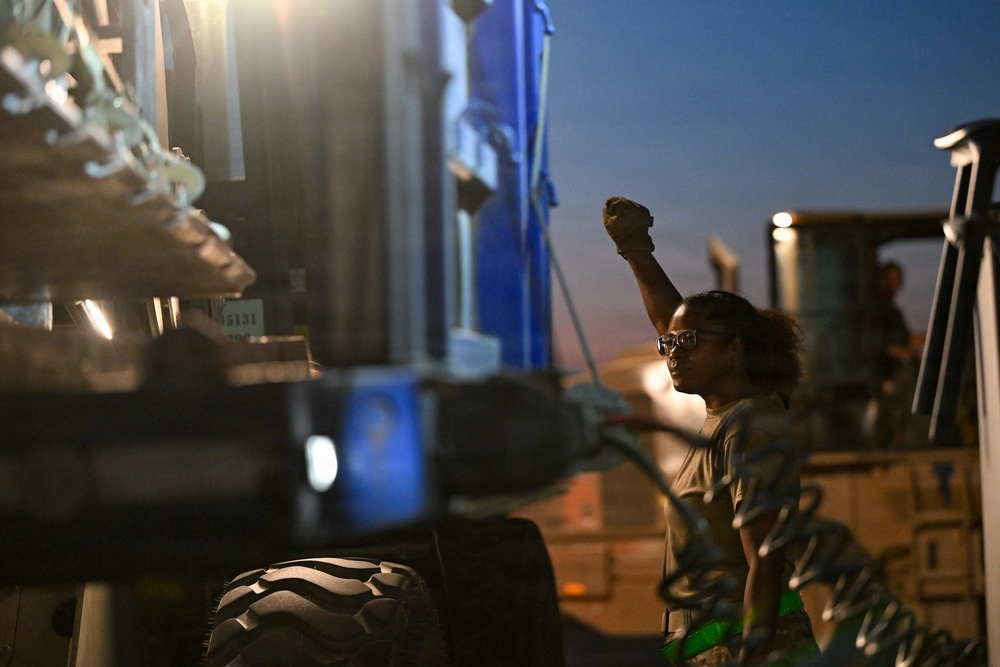 A person with curly hair and glasses raises a clenched fist in the air while standing among large industrial vehicles or machinery at dusk or nighttime. The scene is dramatically lit with artificial lighting against a twilight sky. The raised fist gesture is prominently visible as the focal point of the image.
