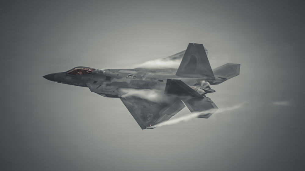 A fifth-generation stealth fighter jet, consistent with the F-22 Raptor design, is captured in flight against an overcast gray sky. The aircraft is performing a high-speed maneuver, evidenced by visible vapor condensation trails forming along the fuselage, wings, and control surfaces. The image is a close-range aerial photograph showcasing the aircraft's angular stealth geometry and dark gray paint scheme.