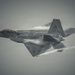 A fifth-generation stealth fighter jet, consistent with the F-22 Raptor design, is captured in flight against an overcast gray sky. The aircraft is performing a high-speed maneuver, evidenced by visible vapor condensation trails forming along the fuselage, wings, and control surfaces. The image is a close-range aerial photograph showcasing the aircraft's angular stealth geometry and dark gray paint scheme.