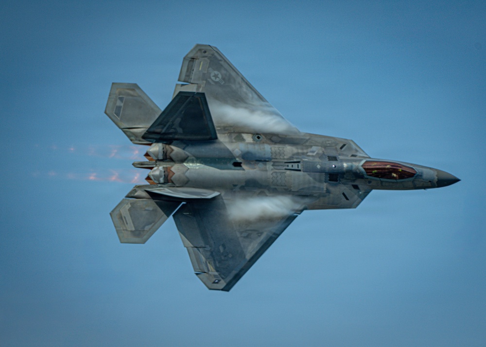 A U.S. Air Force F-22 Raptor fifth-generation stealth fighter jet is captured in a dramatic top-down perspective during a high-performance aerial maneuver. The aircraft is in afterburner with engine exhaust flames visible, and condensation vapor trails are forming over the fuselage due to low-pressure aerodynamic effects. The image showcases the aircraft's distinctive stealth geometry, open weapons bays, and twin-engine configuration.