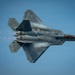 A U.S. Air Force F-22 Raptor fifth-generation stealth fighter jet is captured in a dramatic top-down perspective during a high-performance aerial maneuver. The aircraft is in afterburner with engine exhaust flames visible, and condensation vapor trails are forming over the fuselage due to low-pressure aerodynamic effects. The image showcases the aircraft's distinctive stealth geometry, open weapons bays, and twin-engine configuration.
