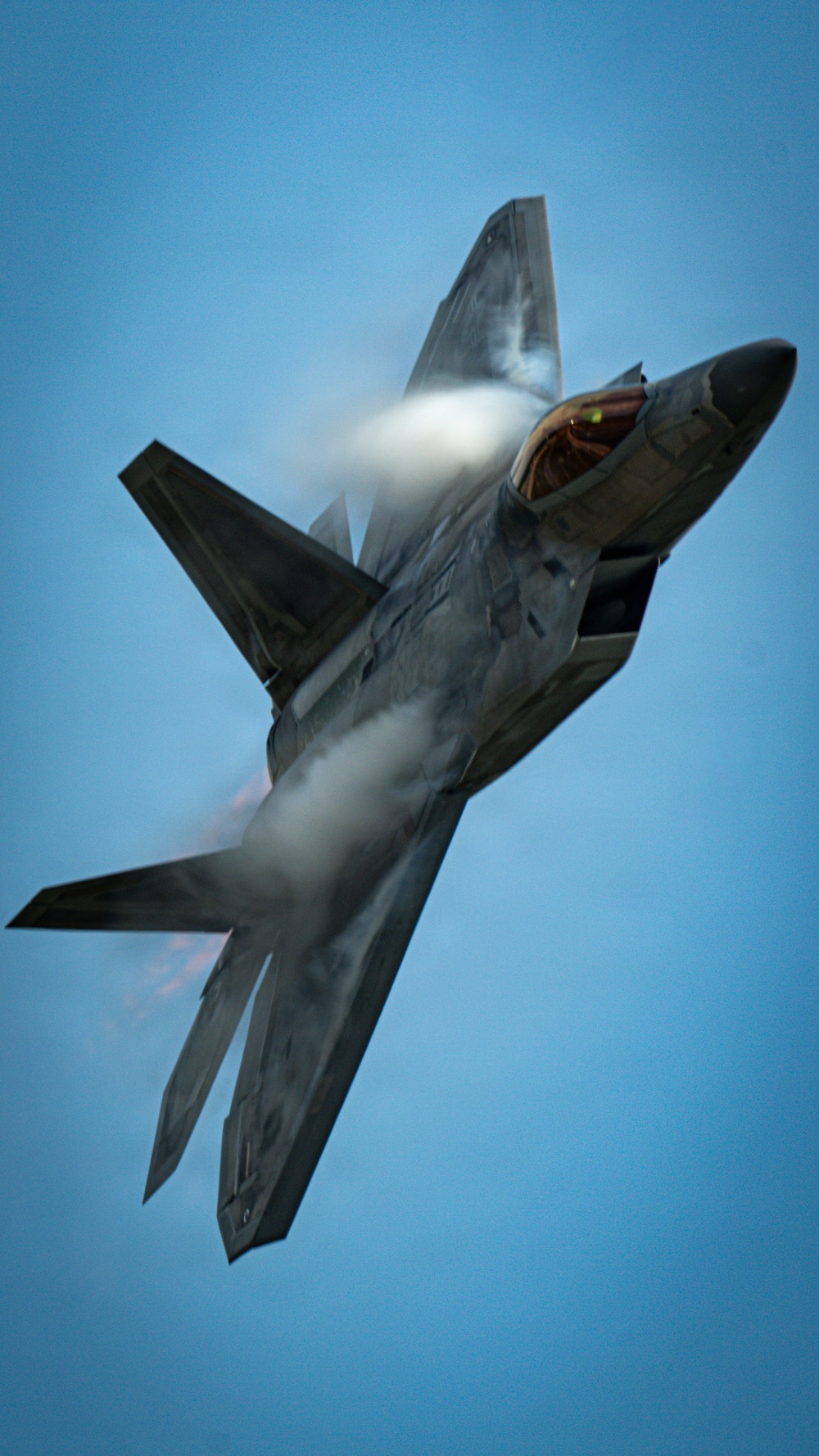 A fifth-generation stealth fighter jet, consistent with an F-22 Raptor, is captured in a steep climbing maneuver against a clear blue sky. The aircraft is generating a dramatic vapor cone or condensation cloud around its fuselage, indicative of high-speed, high-G flight. The cockpit canopy is visible and appears to be open or in a raised position, with interior lighting faintly visible.