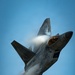 A fifth-generation stealth fighter jet, consistent with an F-22 Raptor, is captured in a steep climbing maneuver against a clear blue sky. The aircraft is generating a dramatic vapor cone or condensation cloud around its fuselage, indicative of high-speed, high-G flight. The cockpit canopy is visible and appears to be open or in a raised position, with interior lighting faintly visible.