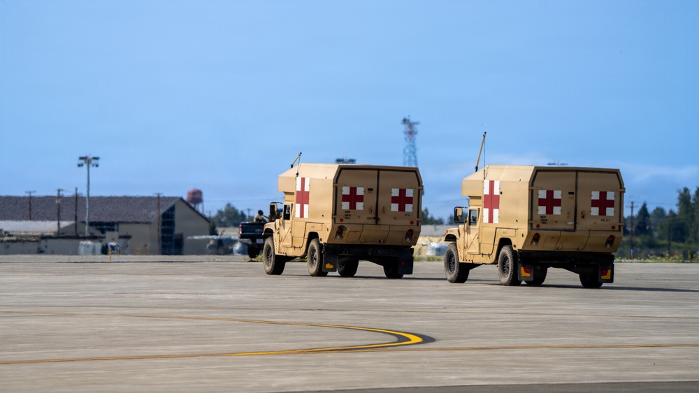 Two military medical HMMWV (Humvee) ambulances are traveling across what appears to be a military airfield tarmac. Both vehicles are painted in desert tan and prominently display red cross markings on their rear panels, identifying them as protected medical vehicles under the Geneva Convention. The setting appears to be a U.S. military installation based on the infrastructure visible in the background.