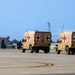 Two military medical HMMWV (Humvee) ambulances are traveling across what appears to be a military airfield tarmac. Both vehicles are painted in desert tan and prominently display red cross markings on their rear panels, identifying them as protected medical vehicles under the Geneva Convention. The setting appears to be a U.S. military installation based on the infrastructure visible in the background.