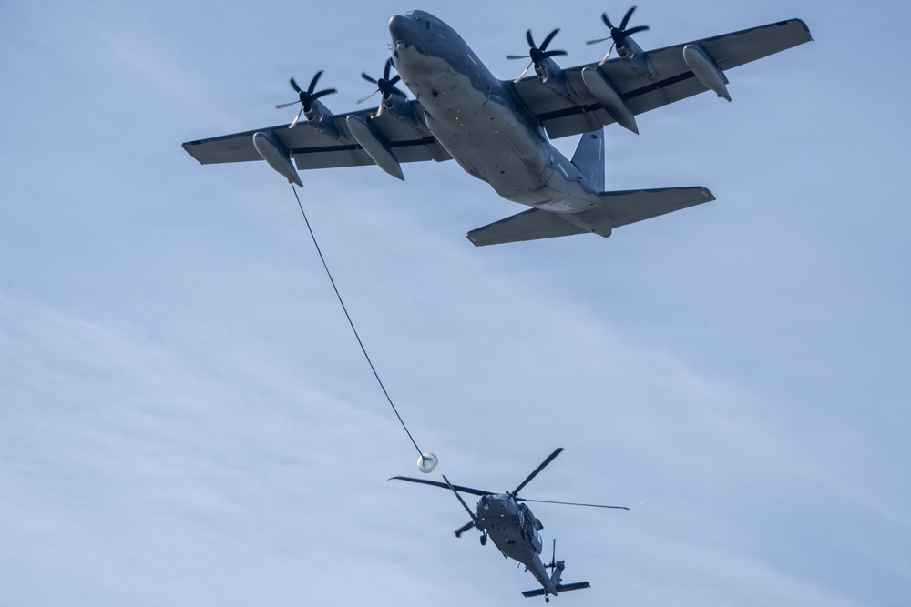 The image shows an aerial refueling operation between a large four-engine turboprop transport aircraft and a military helicopter. The fixed-wing aircraft, consistent with a C-130 variant configured as a tanker, has deployed a hose-and-drogue refueling system toward the helicopter below. The helicopter appears to be approaching or connected to the drogue basket, conducting an airborne fuel transfer.