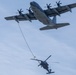 The image shows an aerial refueling operation between a large four-engine turboprop transport aircraft and a military helicopter. The fixed-wing aircraft, consistent with a C-130 variant configured as a tanker, has deployed a hose-and-drogue refueling system toward the helicopter below. The helicopter appears to be approaching or connected to the drogue basket, conducting an airborne fuel transfer.