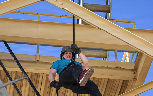 A person is rappelling or abseiling down from a large yellow steel structure, suspended by ropes. They are smiling broadly, suggesting enjoyment or excitement during the activity. The structure appears to be an industrial or recreational tower with staircases and metal framework.