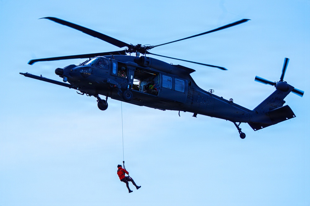A dark military helicopter, consistent with a Sikorsky HH-60 Pave Hawk or UH-60 Black Hawk variant, is hovering against a pale blue sky. A person wearing a bright orange flight suit or rescue suit is suspended below the aircraft on a hoist cable. This appears to be a search and rescue (SAR) operation or training exercise.