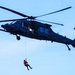 A dark military helicopter, consistent with a Sikorsky HH-60 Pave Hawk or UH-60 Black Hawk variant, is hovering against a pale blue sky. A person wearing a bright orange flight suit or rescue suit is suspended below the aircraft on a hoist cable. This appears to be a search and rescue (SAR) operation or training exercise.