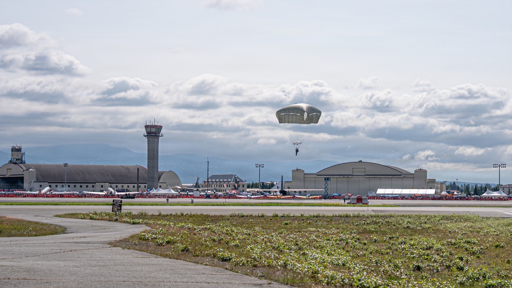 A parachutist descends toward an airfield during what appears to be an air show event, with a large crowd visible along the flight line. The scene includes military or civilian aviation infrastructure including hangars, a control tower, and numerous aircraft on static display. Mountains are faintly visible in the background under a partly cloudy sky.