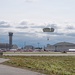 A parachutist descends toward an airfield during what appears to be an air show event, with a large crowd visible along the flight line. The scene includes military or civilian aviation infrastructure including hangars, a control tower, and numerous aircraft on static display. Mountains are faintly visible in the background under a partly cloudy sky.
