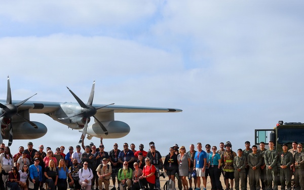 A large group of approximately 60-80 people poses for a group photograph on a military airfield tarmac. The group includes a mix of military personnel in flight suits and civilians in casual attire, gathered in front of a C-130 Hercules transport aircraft bearing the number '26'. A large military vehicle, likely a crash/fire rescue truck, is visible on the right side of the frame.