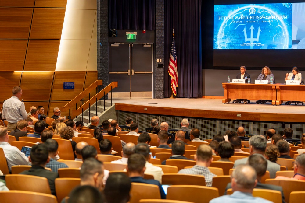 A panel discussion is taking place at the 'Future Warfighting Symposium' with the subtitle 'Innovate | Integrate | Dominate,' held at the United States Naval War College in Newport, Rhode Island, August 3-5, 2025. Three panelists — identified by nameplates as Manchester, Yanez, and Danczak — are seated at a table on stage before a large audience in an auditorium. An American flag stands on stage and a large projection screen displays the event branding.