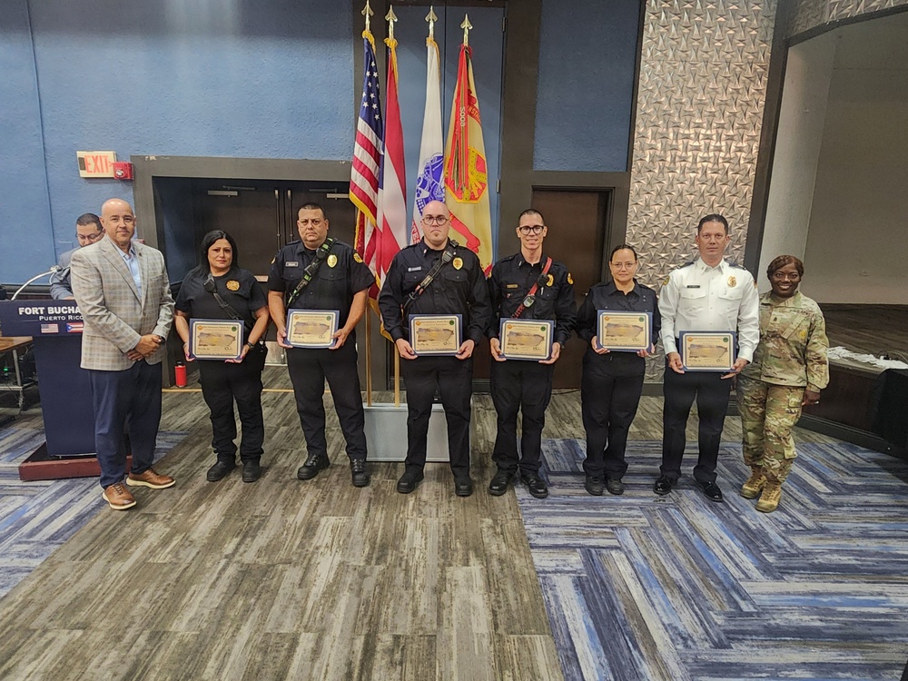A formal awards ceremony taking place at Fort Buchanan, Puerto Rico, where five security or law enforcement personnel in black uniforms are holding certificates of recognition. They are flanked by what appears to be a civilian official on the left, a fire department officer in white uniform, and a military soldier in camouflage on the right. The setting is an official indoor venue with multiple flags including the U.S. flag, Puerto Rico flag, and other organizational flags.