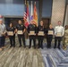 A formal awards ceremony taking place at Fort Buchanan, Puerto Rico, where five security or law enforcement personnel in black uniforms are holding certificates of recognition. They are flanked by what appears to be a civilian official on the left, a fire department officer in white uniform, and a military soldier in camouflage on the right. The setting is an official indoor venue with multiple flags including the U.S. flag, Puerto Rico flag, and other organizational flags.