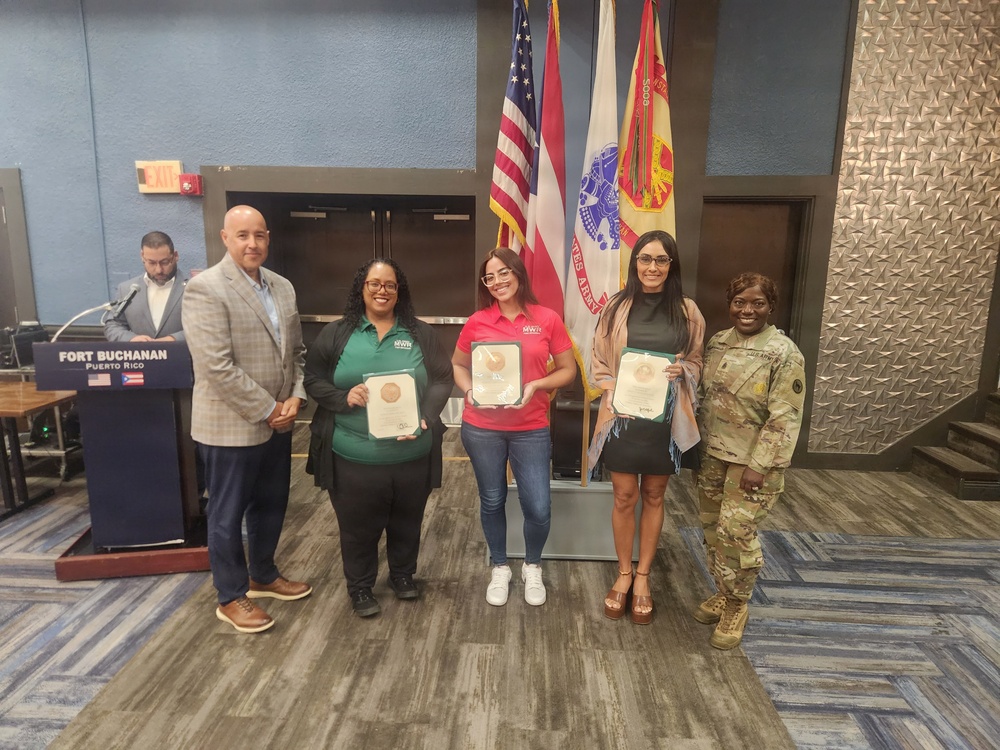A formal recognition ceremony is taking place at Fort Buchanan, Puerto Rico, where three individuals are being presented with awards or certificates. The group poses in front of multiple flags including the American flag, Puerto Rico flag, and U.S. Army flags. A military officer in Army Combat Uniform and a civilian official flank the award recipients.