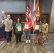 A formal recognition ceremony is taking place at Fort Buchanan, Puerto Rico, where three individuals are being presented with awards or certificates. The group poses in front of multiple flags including the American flag, Puerto Rico flag, and U.S. Army flags. A military officer in Army Combat Uniform and a civilian official flank the award recipients.