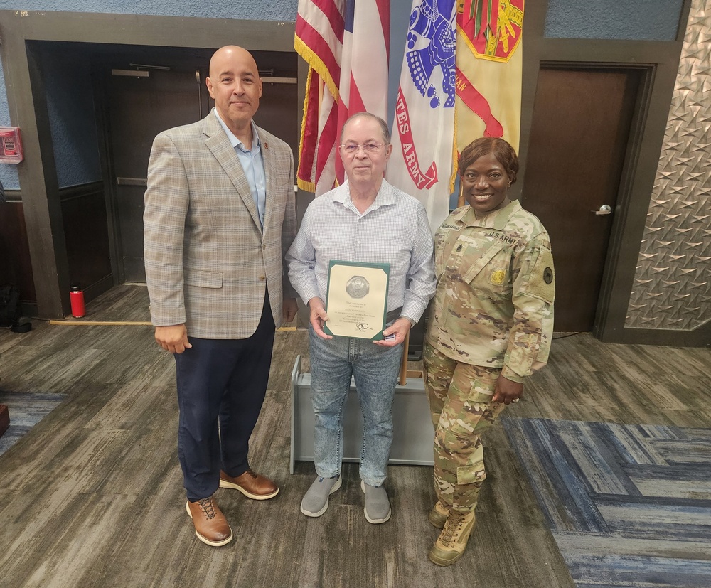 Three individuals pose for a formal photo in what appears to be a U.S. Army facility. The person in the center is holding a framed certificate or award, flanked by a civilian in a blazer on the left and a U.S. Army soldier in MultiCam (OCP) uniform on the right. The setting suggests an official recognition or award ceremony.