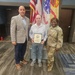 Three individuals pose for a formal photo in what appears to be a U.S. Army facility. The person in the center is holding a framed certificate or award, flanked by a civilian in a blazer on the left and a U.S. Army soldier in MultiCam (OCP) uniform on the right. The setting suggests an official recognition or award ceremony.