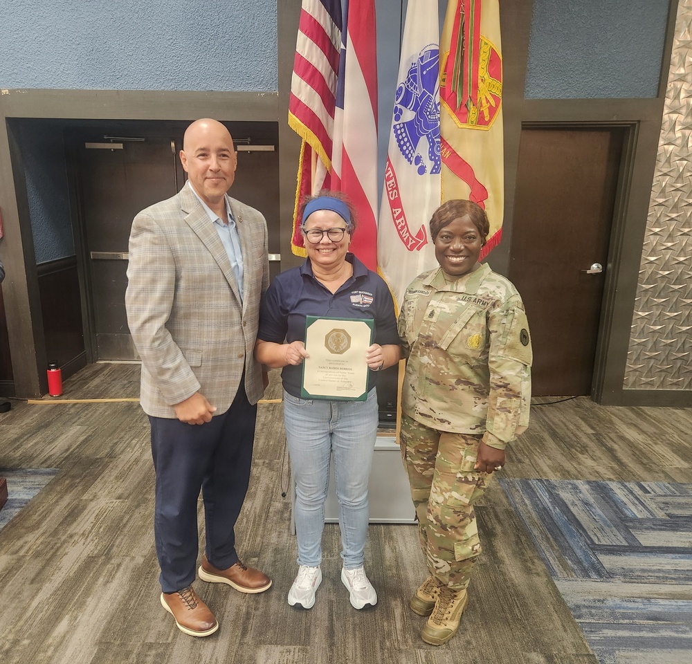 Three individuals pose for a formal photo in what appears to be a U.S. Army facility. The person in the center is holding a certificate or award plaque, flanked by a civilian male in a blazer on the left and a U.S. Army soldier in OCP (Operational Camouflage Pattern) uniform on the right. The setting includes multiple flags including the U.S. flag and a U.S. Army flag in the background.