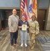 Three individuals pose for a formal photo in what appears to be a U.S. Army facility. The person in the center is holding a certificate or award plaque, flanked by a civilian male in a blazer on the left and a U.S. Army soldier in OCP (Operational Camouflage Pattern) uniform on the right. The setting includes multiple flags including the U.S. flag and a U.S. Army flag in the background.