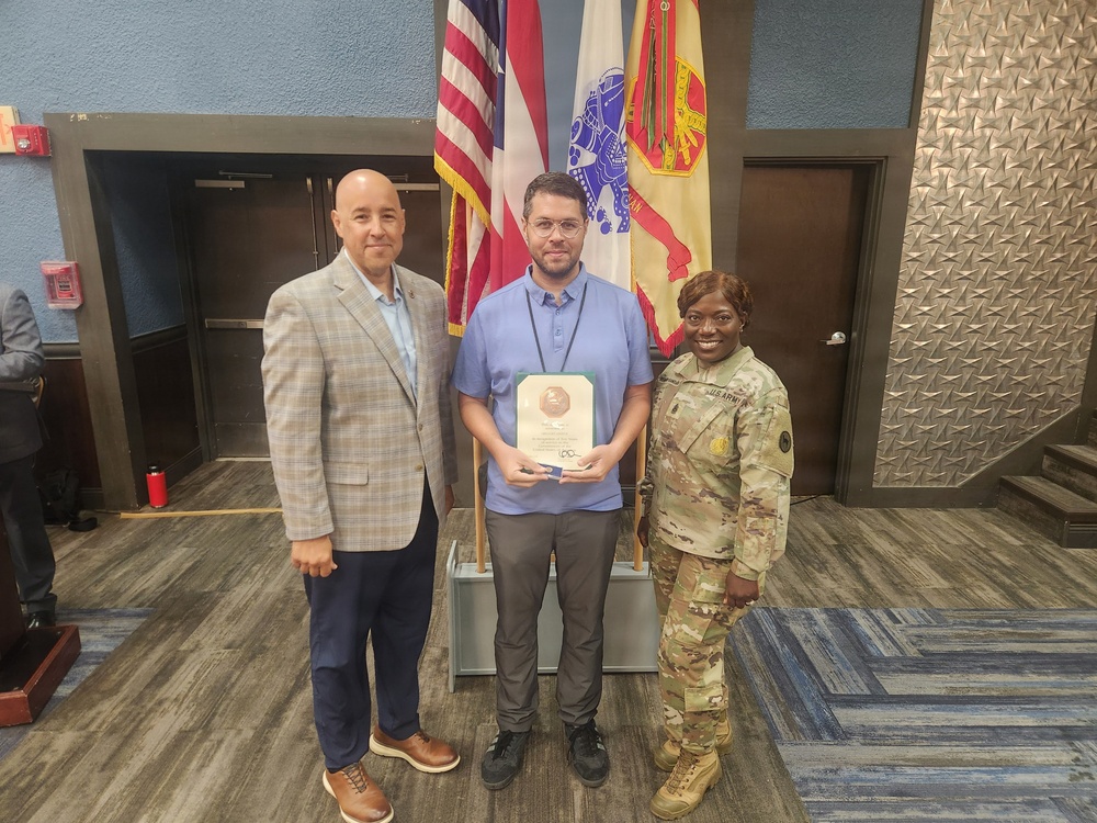 Three individuals are posing for a formal photo in what appears to be a military or government building. The person in the center is holding a framed certificate or award, flanked by a civilian in a blazer on the left and a U.S. Army soldier in camouflage uniform on the right. The setting includes multiple flags including the U.S. flag and what appears to be a U.S. Army flag.