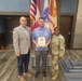 Three individuals are posing for a formal photo in what appears to be a military or government building. The person in the center is holding a framed certificate or award, flanked by a civilian in a blazer on the left and a U.S. Army soldier in camouflage uniform on the right. The setting includes multiple flags including the U.S. flag and what appears to be a U.S. Army flag.