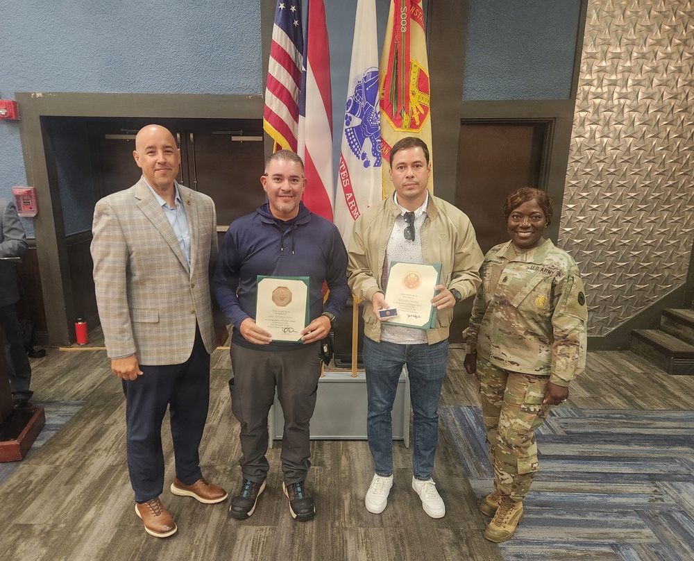 Four individuals pose for a formal group photo in what appears to be a U.S. Army facility. Two civilians in the center are holding official-looking award certificates or commendation plaques with green folders and gold medallions. The group includes a man in a blazer, two civilian recipients, and a U.S. Army soldier in OCP (Operational Camouflage Pattern) uniform.