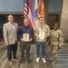 Four individuals pose for a formal group photo in what appears to be a U.S. Army facility. Two civilians in the center are holding official-looking award certificates or commendation plaques with green folders and gold medallions. The group includes a man in a blazer, two civilian recipients, and a U.S. Army soldier in OCP (Operational Camouflage Pattern) uniform.