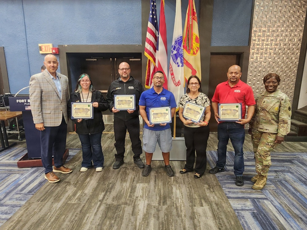 A group of seven people pose for a formal recognition photo at what appears to be a Fort Buchanan military installation. Five individuals are holding certificates or plaques, flanked by a civilian presenter on the left and a U.S. Army soldier in OCP (Operational Camouflage Pattern) uniform on the right. The setting includes U.S. and military flags in the background, suggesting an official awards ceremony.