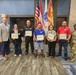 A group of seven people pose for a formal recognition photo at what appears to be a Fort Buchanan military installation. Five individuals are holding certificates or plaques, flanked by a civilian presenter on the left and a U.S. Army soldier in OCP (Operational Camouflage Pattern) uniform on the right. The setting includes U.S. and military flags in the background, suggesting an official awards ceremony.
