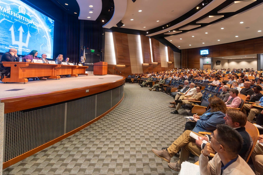 A large professional symposium titled 'Warfighting Symposium' is taking place in a modern auditorium. A panel of approximately five to six speakers is seated at a curved wooden dais on stage, facing a packed audience of hundreds of attendees. The event appears to be affiliated with a U.S. military institution, suggested by the trident logo and naval insignia visible on the screen and seating.