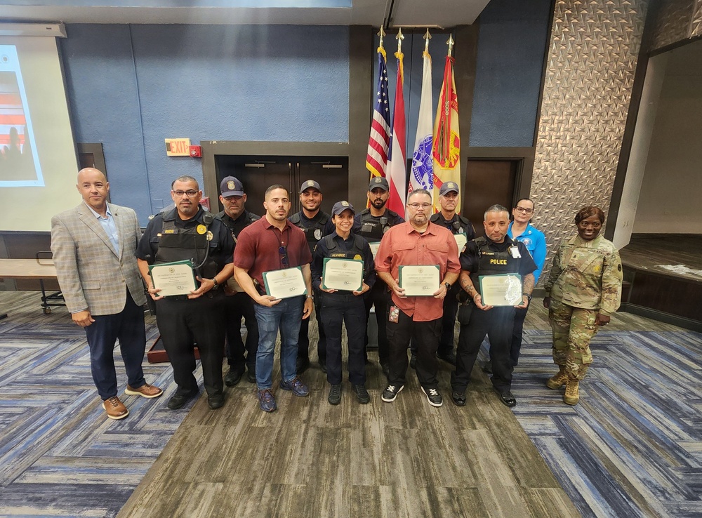 A group of approximately 13 individuals pose for a formal group photo in what appears to be a conference or meeting room. Several participants in the front row are holding certificates, suggesting a recognition or graduation ceremony. The group includes uniformed security/police officers, civilians, a military soldier in camouflage uniform, and a person in a business suit.