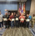 A group of approximately 13 individuals pose for a formal group photo in what appears to be a conference or meeting room. Several participants in the front row are holding certificates, suggesting a recognition or graduation ceremony. The group includes uniformed security/police officers, civilians, a military soldier in camouflage uniform, and a person in a business suit.