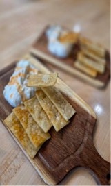 The image shows two wooden serving boards on a light wooden table, each containing thin crispy crackers or flatbread chips alongside what appears to be a soft white cheese, possibly brie or camembert with a golden topping. The foreground board is in sharp focus while the background board is blurred. This appears to be a restaurant or café appetizer presentation.