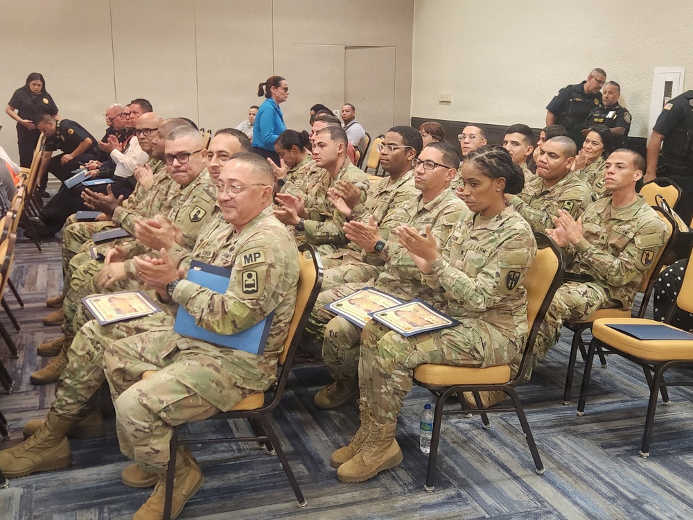 A group of U.S. Army soldiers in OCP (Operational Camouflage Pattern) uniforms are seated in rows of yellow chairs at what appears to be a formal ceremony or award presentation. Many soldiers are holding blue folders containing certificates and are actively applauding. Civilian law enforcement officers in black uniforms are visible in the background.