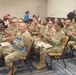 A group of U.S. Army soldiers in OCP (Operational Camouflage Pattern) uniforms are seated in rows of yellow chairs at what appears to be a formal ceremony or award presentation. Many soldiers are holding blue folders containing certificates and are actively applauding. Civilian law enforcement officers in black uniforms are visible in the background.
