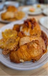 A close-up shot of a plate of food featuring what appears to be fried chicken pieces with a crispy, golden-brown exterior, accompanied by fried plantains and a red bean or tomato-based sauce. The dish appears to be a Latin or Caribbean-style meal served on a white ceramic plate. Additional plates of food are visible in the blurred background, suggesting a restaurant or communal dining setting.