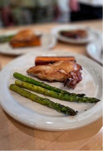 A plated meal on a white ceramic dish featuring roasted or grilled chicken, asparagus spears, and what appears to be roasted carrots. The dish is presented on a light wood table in what appears to be a restaurant setting. Additional plates with food are visible in the blurred background.
