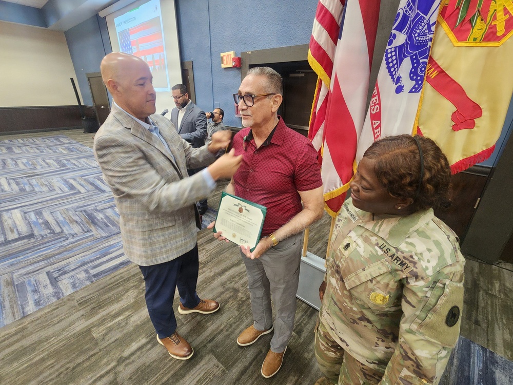 A formal ceremony is taking place in what appears to be a military or government meeting room. A civilian in a plaid blazer is pinning something onto a man in a red shirt who is holding a certificate, while a U.S. Army soldier in camouflage uniform stands nearby. Military flags including the U.S. flag and an Army unit flag are displayed in the background.