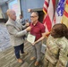 A formal ceremony is taking place in what appears to be a military or government meeting room. A civilian in a plaid blazer is pinning something onto a man in a red shirt who is holding a certificate, while a U.S. Army soldier in camouflage uniform stands nearby. Military flags including the U.S. flag and an Army unit flag are displayed in the background.