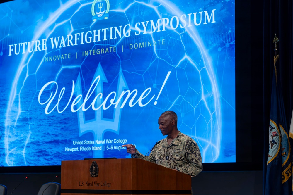 A U.S. Navy officer in camouflage uniform is speaking at a podium during the Future Warfighting Symposium at the U.S. Naval War College in Newport, Rhode Island. The event, themed 'Innovate | Integrate | Dominate,' took place on 5-6 August. A large projection screen behind the speaker displays a 'Welcome!' slide with the symposium branding.