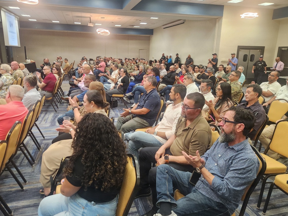 A large indoor gathering or meeting is taking place in a conference or banquet hall with rows of gold/yellow chairs filled with attendees. The crowd appears to be watching a presentation, indicated by a projection screen visible on the left side. Security personnel in black uniforms are visible standing along the back wall.