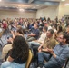 A large indoor gathering or meeting is taking place in a conference or banquet hall with rows of gold/yellow chairs filled with attendees. The crowd appears to be watching a presentation, indicated by a projection screen visible on the left side. Security personnel in black uniforms are visible standing along the back wall.