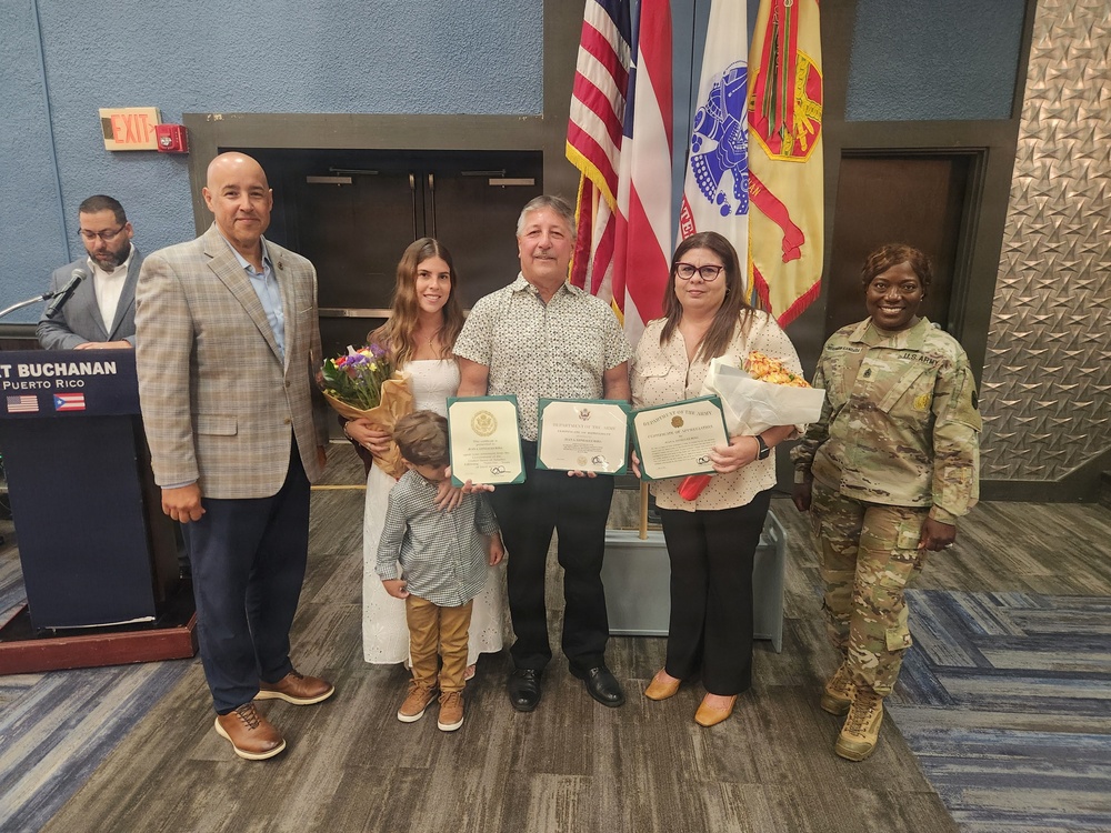 A formal award ceremony is taking place at Fort Buchanan in Puerto Rico, where civilians are being recognized with framed certificates. A group of approximately six adults and one child pose for a photo, with the central figure holding multiple award certificates. A U.S. Army soldier in camouflage uniform stands to the right, and flags including the U.S. flag and military unit flags are visible in the background.