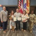 A formal award ceremony is taking place at Fort Buchanan in Puerto Rico, where civilians are being recognized with framed certificates. A group of approximately six adults and one child pose for a photo, with the central figure holding multiple award certificates. A U.S. Army soldier in camouflage uniform stands to the right, and flags including the U.S. flag and military unit flags are visible in the background.